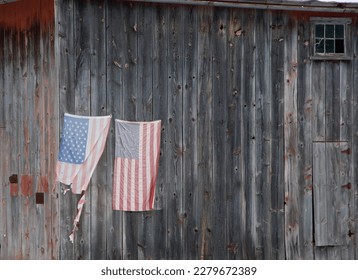 two American flags on the side of an old wood barn wall, with one ripped and torn from the wind