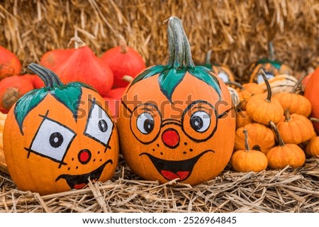 Two amusing painted pumpkins with funny faces, surrounded by smaller pumpkins on a straw background, adding a playful Halloween atmosphere.