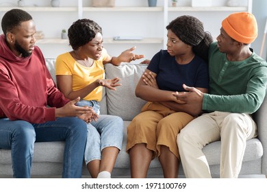 Two Angry Black Girlfriends Fighting While Having Double Date At Home, African American Guys Hugging Them, Sitting On Couch All Together In Living Room. Friends Having Quarrel