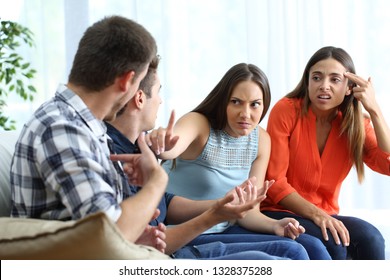 Two Angry Men And Women Arguing Sitting On A Couch In The Living Room At Home