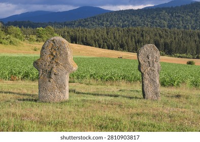 Two anthropomorphic stone monuments at Maculje Necropolis near Novi Travnik, Bosnia and Herzegovina, standing above cornfields and a forested mountain landscape.