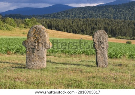 Two anthropomorphic stone monuments at Maculje Necropolis near Novi Travnik, Bosnia and Herzegovina, standing above cornfields and a forested mountain landscape.