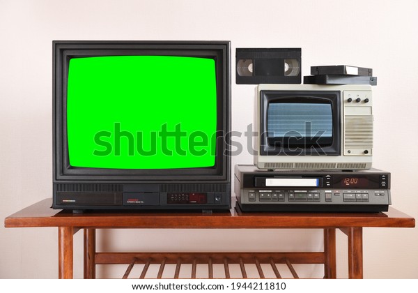 Two antiquated vintage TVs with VCR with green\
screens and noisy screens sit on a vintage table in a tenement\
house in the 1990s, 1980s.