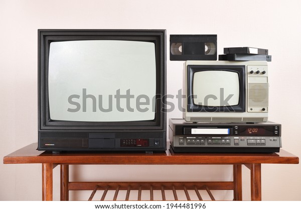 Two antiquated
vintage TVs with VCRs sit on a vintage table in a tenement building
from the 1990s, 1980s.