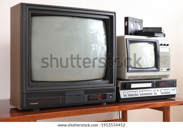 Two antiquated\
vintage TVs with VCRs sit on a vintage table in a tenement building\
from the 1990s, 1980s.