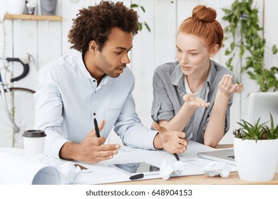 Two architect workers sitting at their cozy workplace surrounded with many papers and modern electronic devices discussing new construction project with emotions using gestures and body language