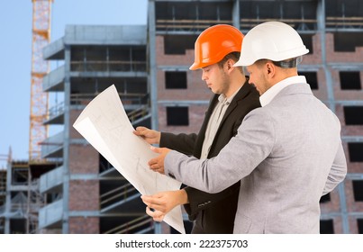 Two architects or engineers standing discussing a building blueprint as they stand on a building site in front of a half completed high-rise building