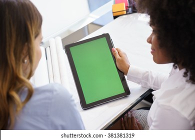 Two architects sitting on desk with blueprints and housing projects. The women hold a tablet and surf the web. The display has a green screen