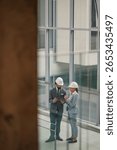 Two architects wearing hardhats are using a tablet and discussing project plans in a modern office building