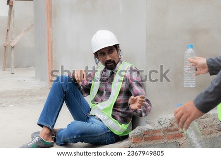 Two Asian builder workers wearing safety reflected vest and helmet tired thirsty hydrated drinking water from plastic bottle together relaxed sitting on construction site on overheated sunny day