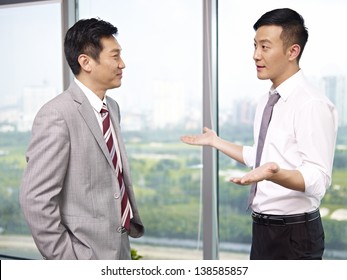 Two Asian Business Executives Standing And Talking By The Windows In Office.