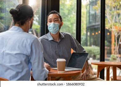 Two Asian Businessman Wearing Protective Face Mask Talking And Discussing In Coffee Shop Together. Hipster Guys Having Friendly Conversation About Business Information In Café During Covid-19 Pandemic