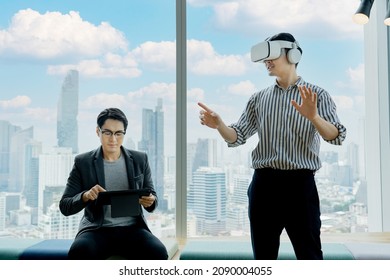 Two Asian businessmen relaxing by one using a tablet, another one playing in a virtual reality simulator headset, in front of a large glass window with a blurred scenery of buildings as background. - Powered by Shutterstock