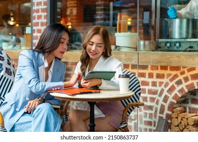 Two Asian businesswoman colleague discussion business plan while sitting at outdoor coffee shop urban city street. Happy business woman with using digital tablet working corporate teamwork together - Powered by Shutterstock
