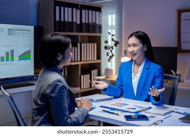 Two asian businesswomen are discussing work in the office at night, brainstorming and strategizing using data visualization - Powered by Shutterstock