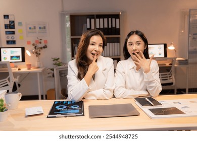 Two asian businesswomen whispering at office desk late at night, sharing secrets and looking surprised. Glow of laptop screen illuminates their faces as they discuss work documents - Powered by Shutterstock