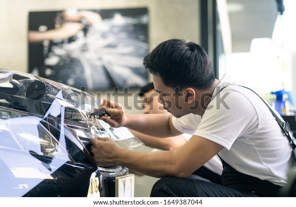 Two Asian car film protection male worker\
installing film to black color car in garage. Man focus holding\
film carefully and cutting film in shape. Coating and protective\
for vehicle business\
concept.
