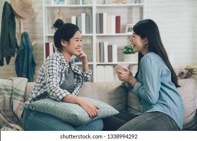 Two Asian Casual Women Relaxing On Sofa With Hot Drink In New Home. Young Girls Chatting Talking About Funny Things Gossip Laughing On Couch In Apartment. Female Roommates Stay In Dormitory Together.