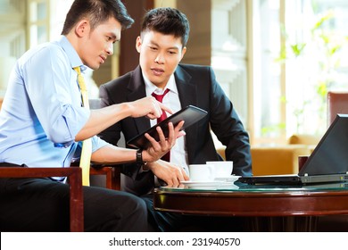 Two Asian Chinese Businessman Or Office People Having A Business Meeting In A Hotel Lobby Discussing Documents On A Tablet Computer While Drinking Coffee 