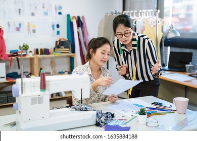 Two Asian Chinese Women Meeting In Fashion Design Studio. Female Creative Dressmakers Talking About Plan On Sketch Paper At Garment Factory. Lady Colleagues Discussing Design Of New Dress On Painting