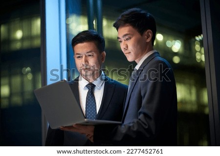 two asian corporate executives standing by the window in office discussing business using laptop computer at night