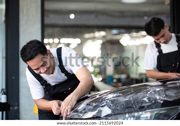 Two Asian detailing film paint protection men\
specialist cutting and wrapping car in garage showroom, protective\
coating installation and polishing automobile for vehicle care\
service business concept
