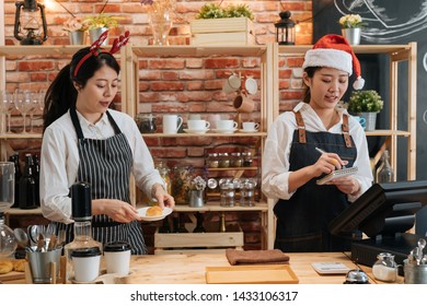 two asian female bartenders dressed in santa hat and working in bar counter in christmas holidays. woman barista prepare croissant while coworker waitress taking note on customer order in xmas. - Powered by Shutterstock