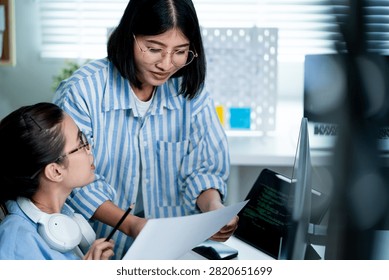Two Asian female software developers discuss a programming project and review computer work together in a modern technology workplace. Their collaborative interaction represents software development, coding, technical communication, teamwork, project management, digital innovation, and professional women working in information technology.