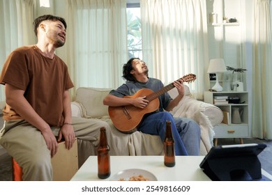 Two Asian friends enjoying a casual music session with acoustic guitar in a living room. Bearded male playing the guitar and the other listening with eyes closed while plays cajon, relaxed ambiance - Powered by Shutterstock