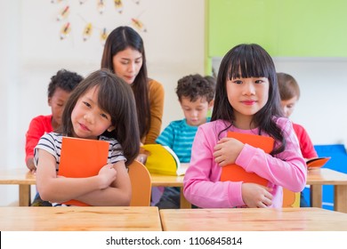 Two Asian Girl Kid Hugging Book And Smiling In Classroom And While Teacher Teach Friends Beside Them,kindergarten Education.