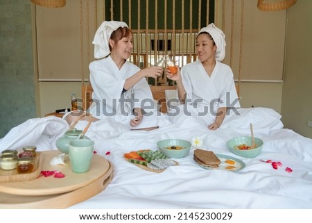 Two Asian girls wearing white bathrobes sit on the bed, eat breakfast together ,drink orange juice together and have fruit, bread, black eggs, tea set, having fun chatting in the hotel.