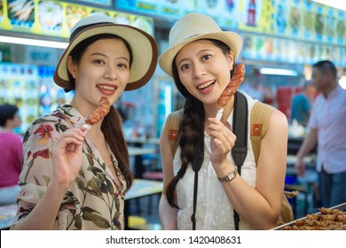Two asian japanese girls friends traveler in taiwan taipei having fun and eating fried sausage at fair market. local food festival concept. young female sisters tourists looking camera face smiling.