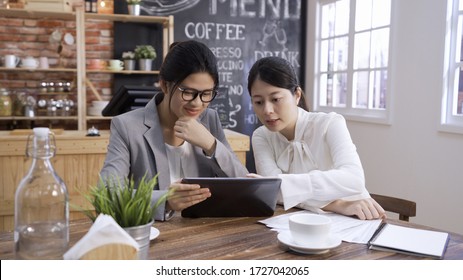 Two Asian Korean Businesswomen Sitting In Coffee House Discussing Business Using Digital Tablet. Elegant Female Worker Colleagues Meeting In Cafe Bar Indoors. Young Employees Talking On Touch Pad