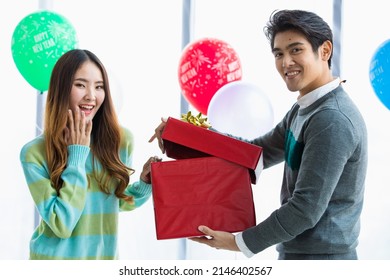 Two Asian lovers enjoying gift exchange together with happy faces at romantic indoor party decorated by fancy balloon to celebrate ecstatic love in winter