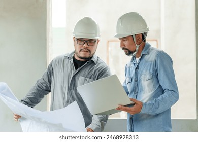 Two Asian male engineers with hard hats reviewing blueprints and laptop at construction site. Concrete wall background. Collaborative and detailed atmosphere. Project planning and execution concept. - Powered by Shutterstock