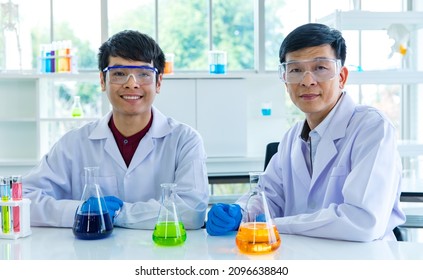 Two Asian male scientists in white lab coat safety goggles rubber gloves working discussing talking together about colorful liquid samples in glass flasks in scientific experiment on laboratory table. - Powered by Shutterstock