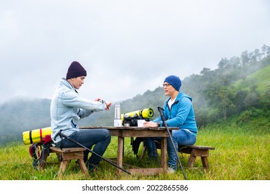 Two Asian Man Enjoy Hiking Together On Forest Mountain In Winter Day. Happy Friends Backpacker Sitting On Outdoor Chair With Drinking Hot Water. Friendship And Outdoors Holiday Vacation Concept.