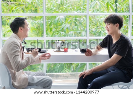Two Asian men are talking  at window with green background in coffee shop.