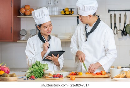Two Asian professional couple chef wearing white uniform, hat, preparing ingredients, cooking meal in kitchen together, using tablet for recipe, smiling with happiness and confidence. Food Concept