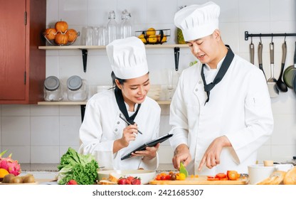 Two Asian professional couple chef wearing white uniform, hat, preparing ingredients, cooking meal in kitchen together, using tablet for recipe, smiling with happiness and confidence. Food Concept - Powered by Shutterstock