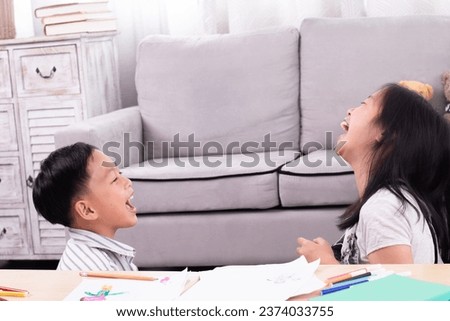 Two Asian siblings boy and girls having fun laughing and drawing cartoons on paper in living room on holiday at home, family relationship childhood lifestyle playing together funny naughty kids health