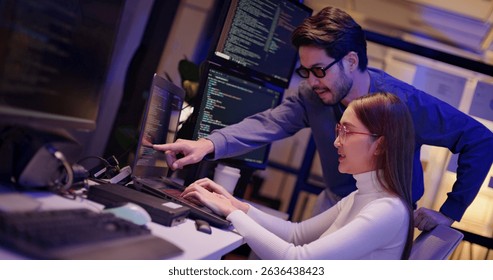 Two Asian software engineers collaborate at night in modern office, writing machine learning algorithms and debugging artificial intelligence code on multiple monitors in AI tech development workflow.