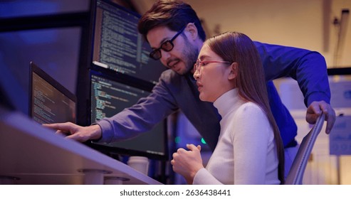 Two Asian software engineers collaborate at night in modern office, writing machine learning algorithms and debugging artificial intelligence code on multiple monitors in AI tech development workflow.