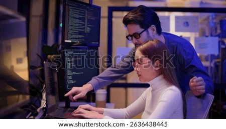 Two Asian software engineers collaborate at night in modern office, writing machine learning algorithms and debugging artificial intelligence code on multiple monitors in AI tech development workflow.