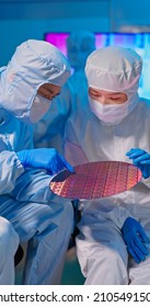 Two Asian Technicians In Sterile Coverall Hold Wafer With Gloves That Reflects Many Different Colors And Check It At Semiconductor Manufacturing Plant