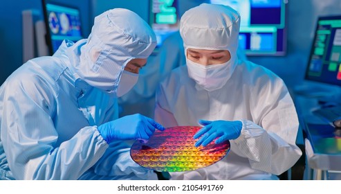 two asian technicians in sterile coverall hold wafer with gloves that reflects many different colors and check it at semiconductor manufacturing plant - Shutterstock ID 2105491769