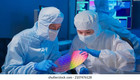 Two Asian Technicians In Sterile Coverall Hold Wafer With Gloves That Reflects Many Different Colors And Check It At Semiconductor Manufacturing Plant