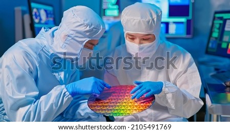 two asian technicians in sterile coverall hold wafer with gloves that reflects many different colors and check it at semiconductor manufacturing plant