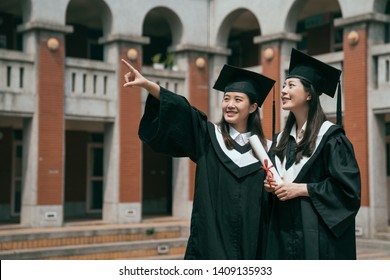 Two Asian Woman College Graduate With Diploma Pointing Up To Sky Looking With Confident Smile. Future Hope Achievement Trend Work Job Seeker. Girls On Graduation Day Standing Outdoor Red Brick Wall