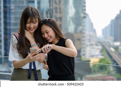 Two Asian Woman Friends Using Mobile Phone At Rooftop With Skyscrapers In City

